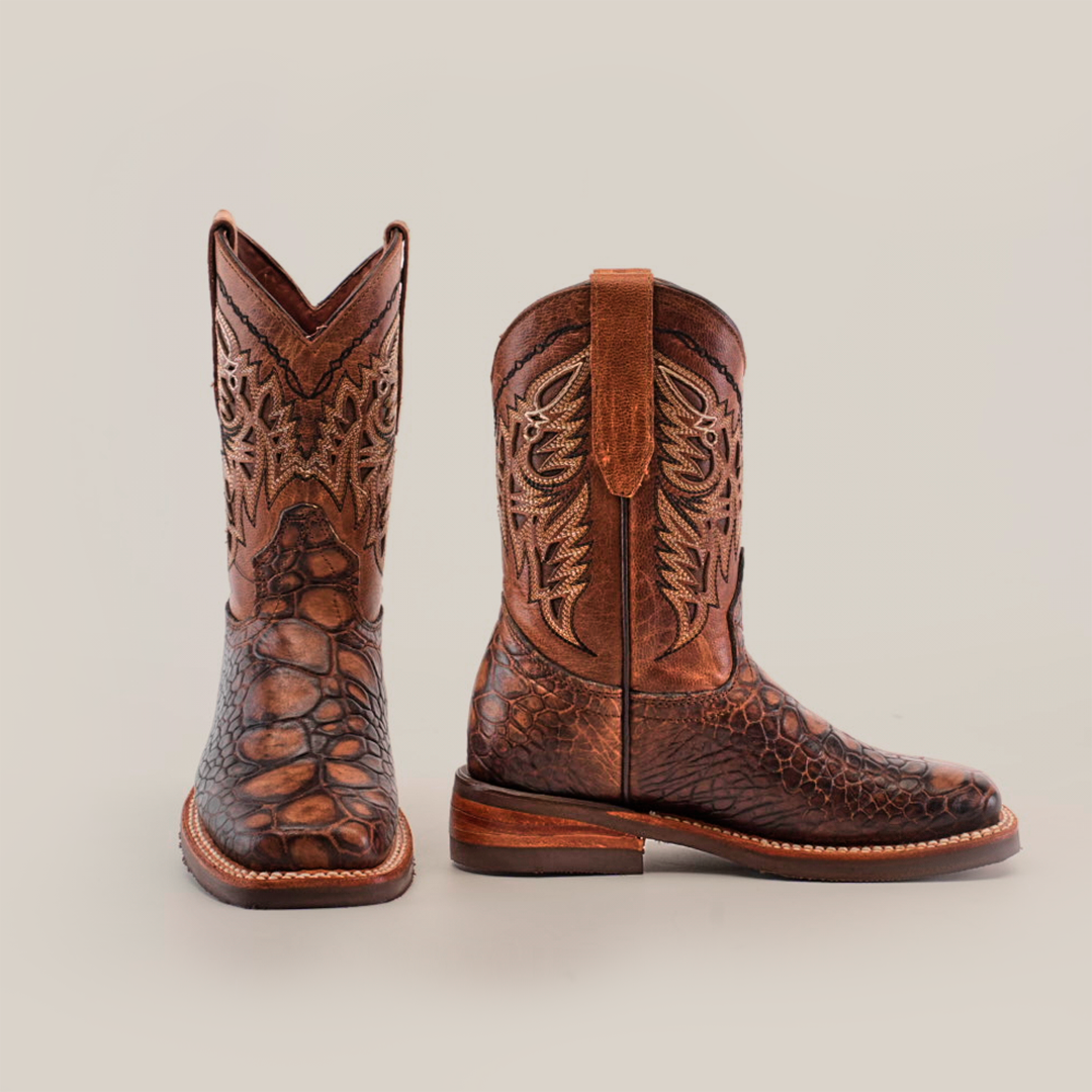 The Loggerhead Turtle Print brown Rodeo Toe boots are crafted from premium cowhide leather, featuring intricate stitching and a scaly texture, reminiscent of loggerhead turtle print. These childrens cowboy boots have a flat heel and are showcased against a plain white background.