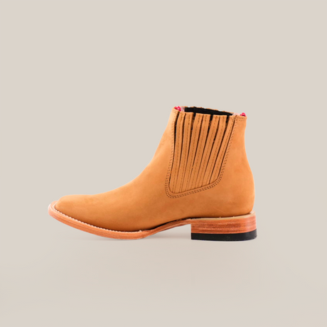 The Prime Suede Honey Square Toe is a tan leather Chelsea boot featuring a wooden heel and black rubber sole, with a hint of cowboy style. It has elastic side panels for easy slip-on and a square toe for a modern look, set against a plain white background.