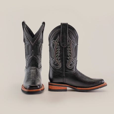 The Barcelona Black Slip Resistant Sole Rodeo Toe boots are displayed on a white background, with one boot facing forward to show off the ornate stitching and the other side-facing to highlight the wooden heel and detailed design. Theyre crafted from premium leather.