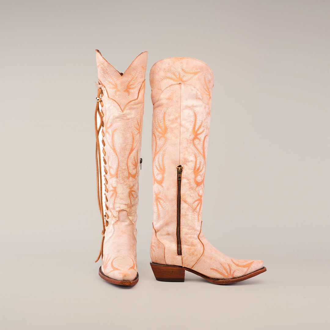 The Emma Alaska Orix-Tall Shaft boots are handcrafted tall tan leather cowboy boots with a subtle antler pattern, featuring a side zipper and heel on one upright boot and decorative lacing on the other against a plain white backdrop.