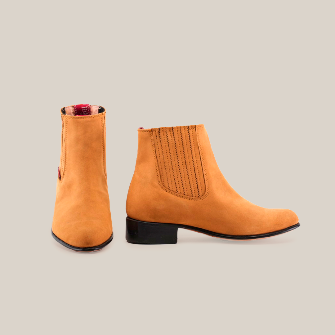 The Prime Suede Honey Round Toe boots feature tan suede, elastic side panels, black low heels, and round toes. One boot shows detailed cowboy-style stitching from the side; the other is front-facing. They have a red and plaid interior lining.