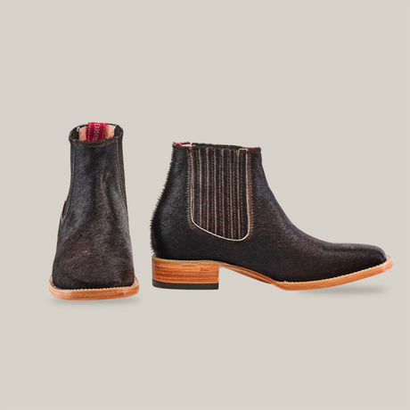 The Cowhide Hair Black Square Toe boots, crafted from cowhide leather with stacked wooden heels, feature elastic side panels and a natural texture. The left boot is positioned forward and the right sideways on a neutral backdrop.