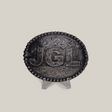 The JGL Buckle, an intricately designed oval piece made from alpaca silver, showcases bold JGL letters and ornate swirling patterns like Texas-style buckles. It is displayed on a clear stand against a plain backdrop.
