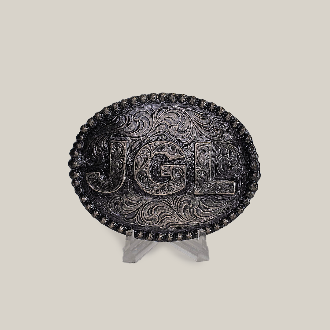 The JGL Buckle, an intricately designed oval piece made from alpaca silver, showcases bold JGL letters and ornate swirling patterns like Texas-style buckles. It is displayed on a clear stand against a plain backdrop.