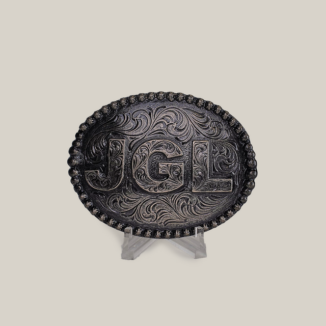 The JGL Buckle, an intricately designed oval piece made from alpaca silver, showcases bold JGL letters and ornate swirling patterns like Texas-style buckles. It is displayed on a clear stand against a plain backdrop.