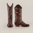 The Dariela Brown Mid Shaft boots, with a narrow square toe, exemplify artisanal excellence in brown leather, adorned with intricate golden embroidery and floral patterns. The left boot stands upright, revealing handcrafted detailing on a neutral background.